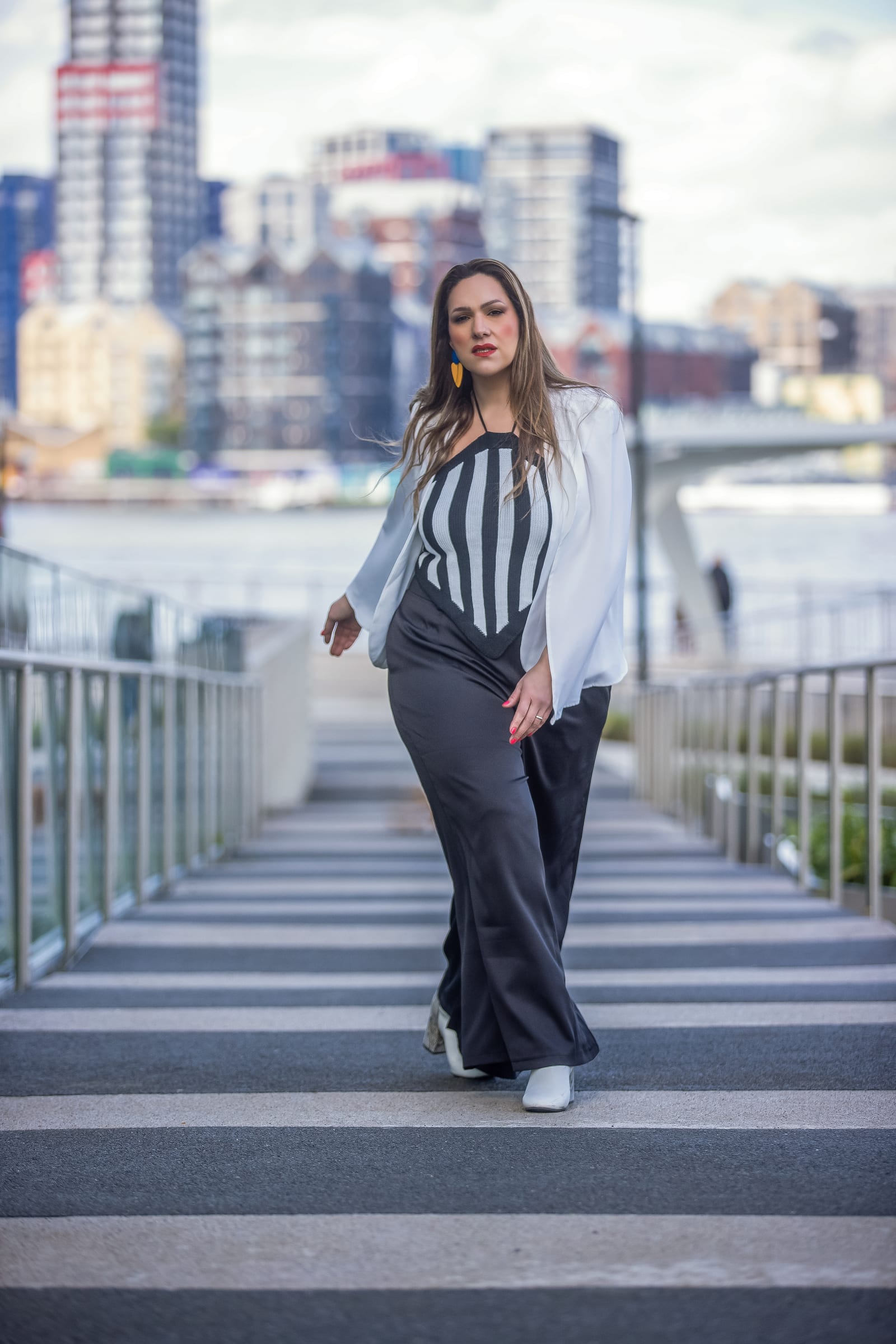 A Ms Curvaceous model walking a riverside walkway in a striped corset and white blazer, the London skyline behind her.